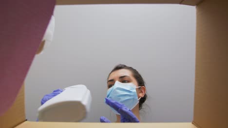 Mujer-Con-Mascarilla-Sacando-Productos-De-Limpieza-De-Una-Caja.-Entrega-A-Domicilio,-Envío-Y-Concepto-De-Pandemia.-Mujer-Con-Mascarilla-Médica-Protectora-Y-Guantes-Abriendo-Una-Caja-De-Paquetería,-Mirando-Dentro-Y-Sacando-Productos-De-Limpieza.